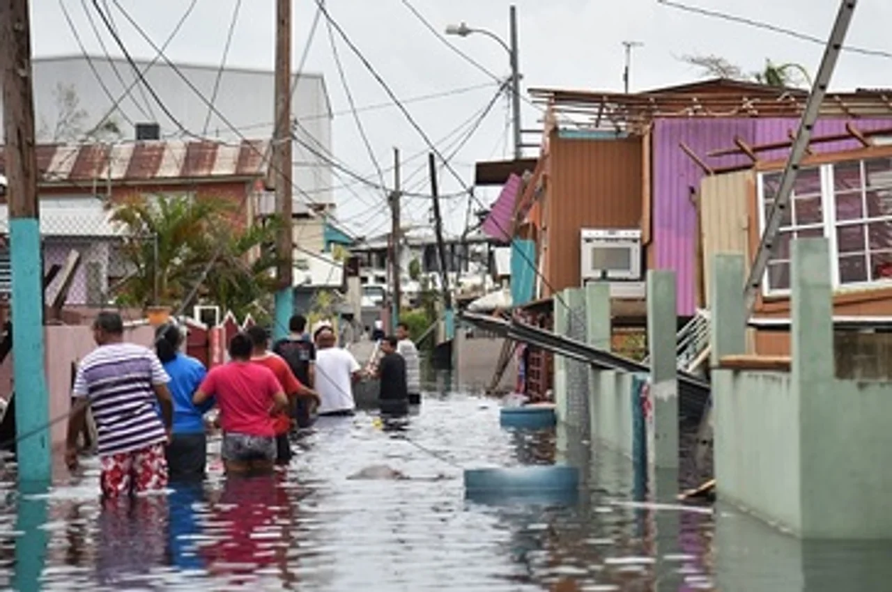 People walk in a flooded street next to damaged houses in Puerto Rico, where Hurricane Maria has created a humanitarian crisis. Photo credit: HECTOR RETAMAL/AFP/Getty Images